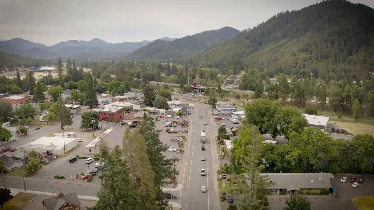 Aerial downtown view of a Southern Oregon small town showing the main street, buildings, parking, and surrounding forested hills.