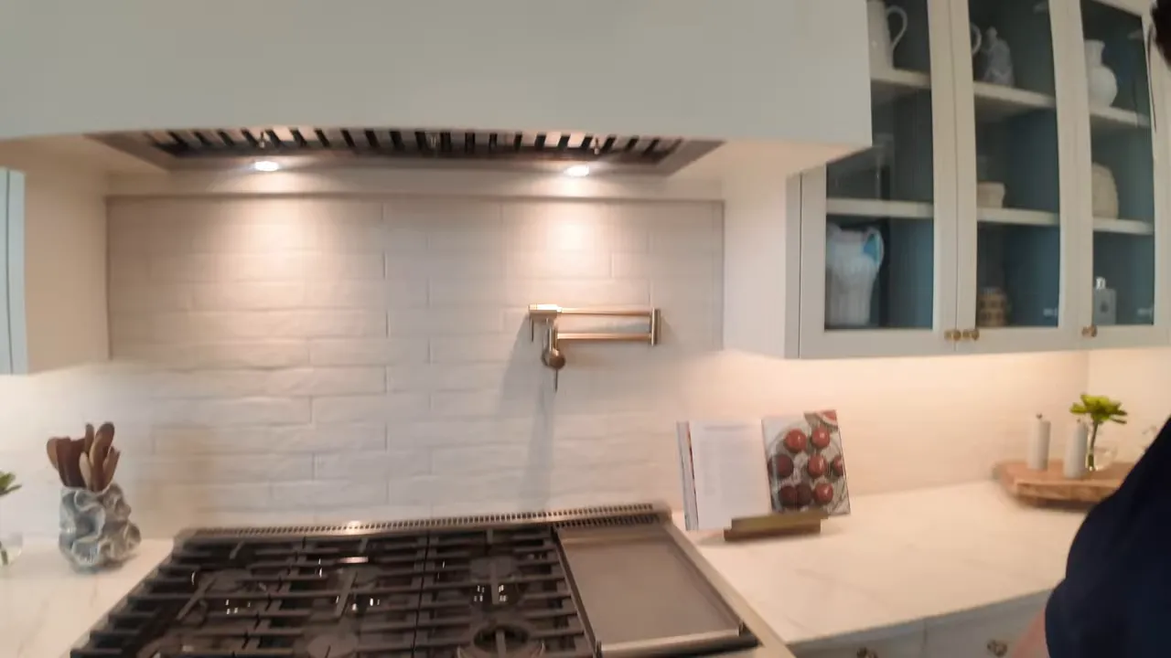 High-end gas range with griddle, textured white tile backsplash, brass pot-filler and glass-front cabinets