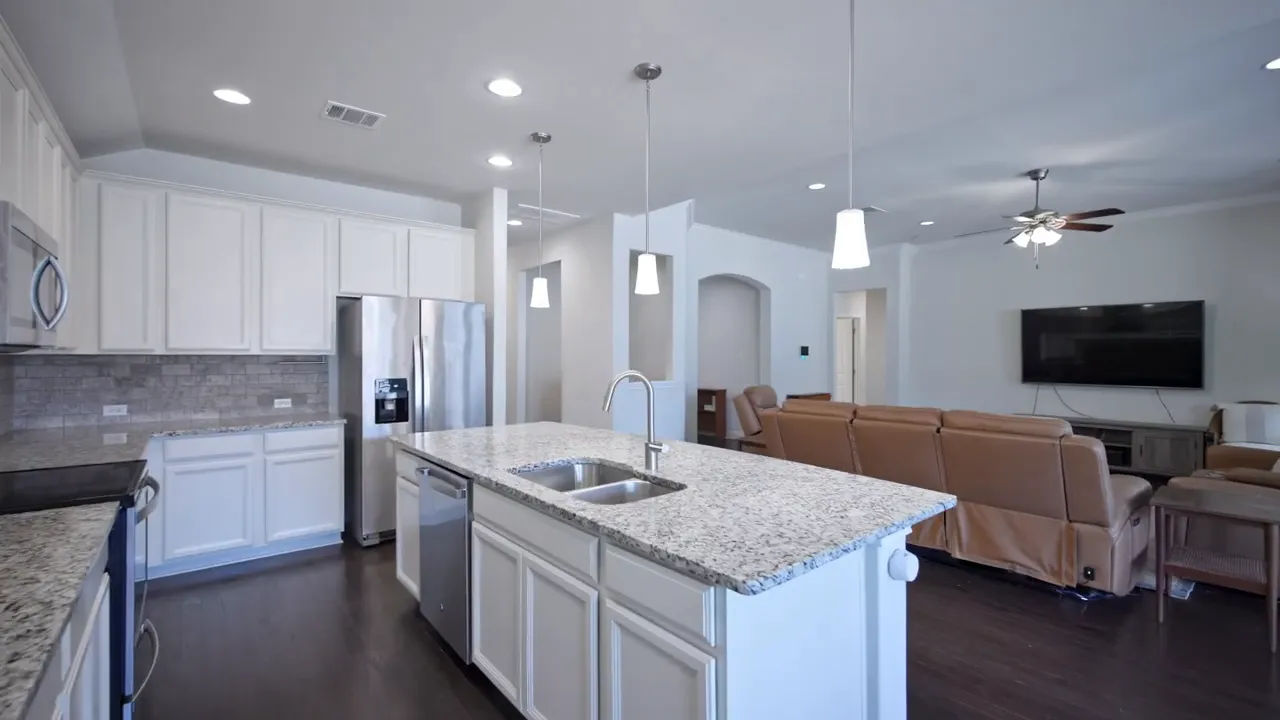 Wide view of kitchen island, white cabinetry, stainless fridge and the adjacent living room with seating and TV