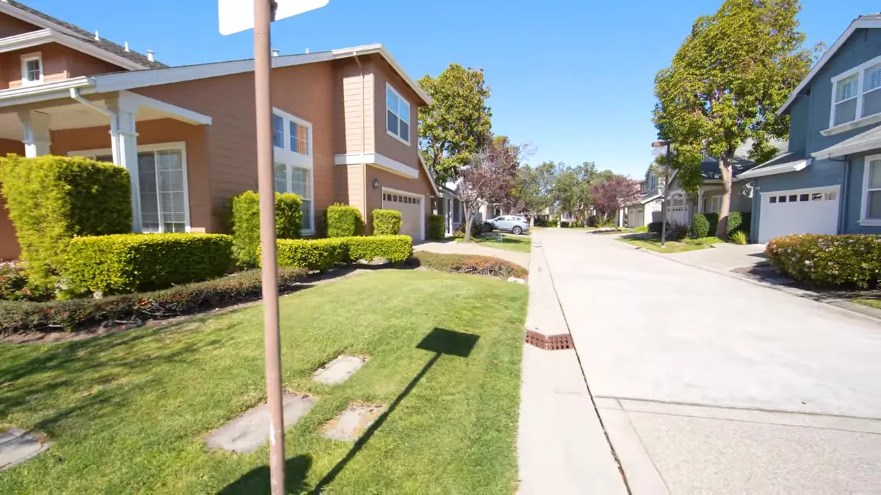Redwood Shores residential street with sidewalks, lawns, manicured hedges and two-story homes under a blue sky.