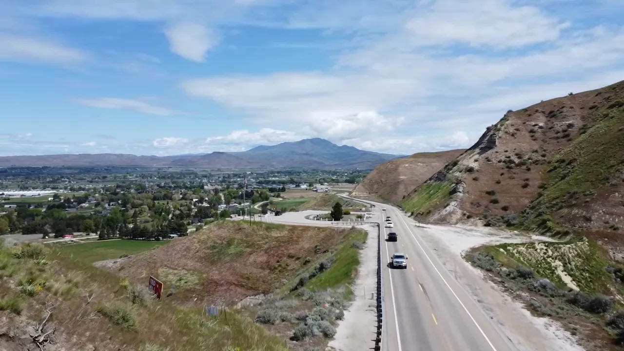 High-angle aerial view of the road descending into Emmett with the town in the valley and mountains beyond under a wide sky.