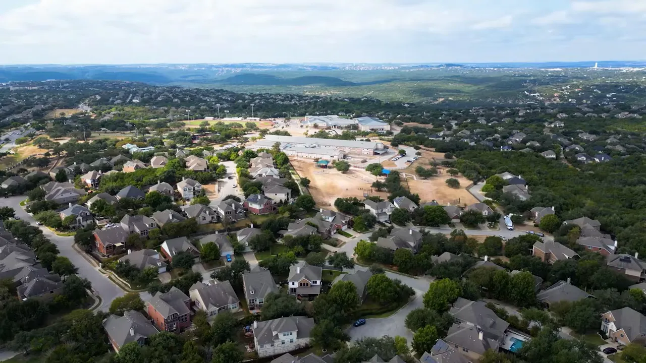 Aerial drone view of a hill-country neighborhood with houses, park/open fields and distant ridgelines