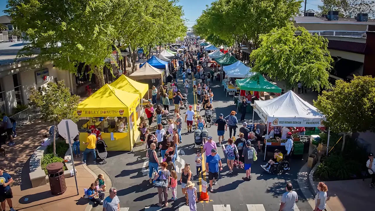 A lively Brentwood street market crowd walking between tents under green trees