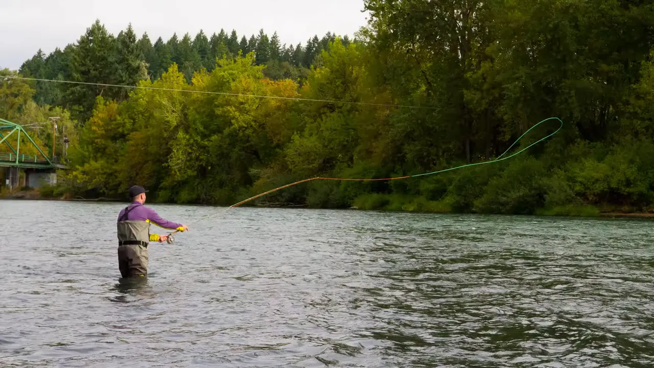 person fly fishing while standing in a river with trees along the bank