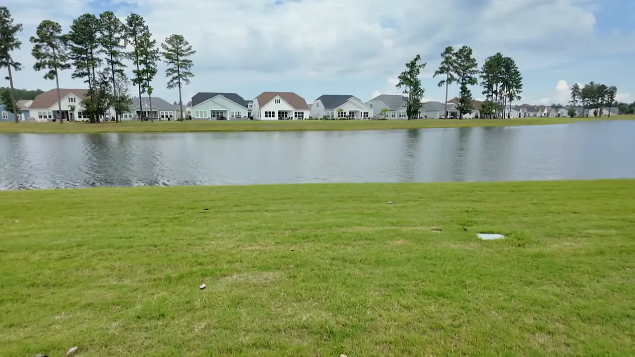 wide lake view with grassy shoreline in the foreground and a row of model homes across the water