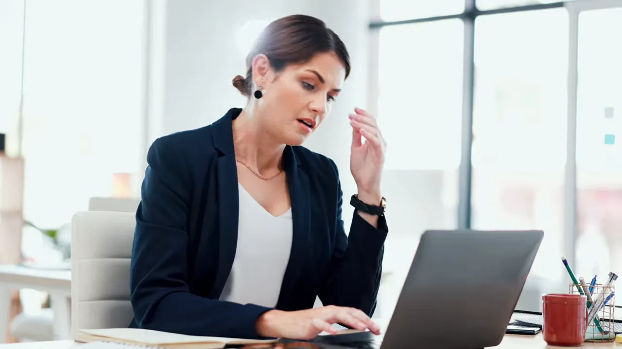 Professional at a laptop appearing stressed and overwhelmed in a bright office