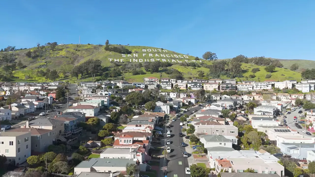 Aerial view of houses below a grassy hillside with large white lettering, illustrating hillside topography vs. flat streets