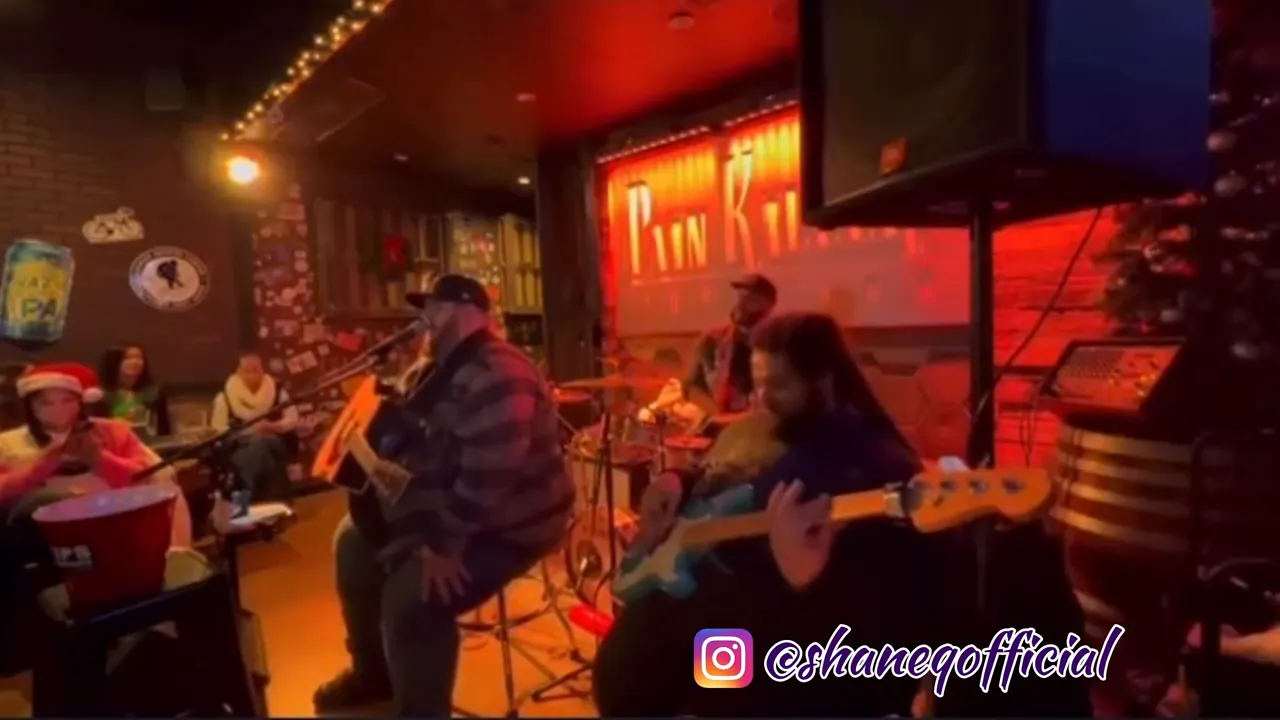 Live band playing in a small club with warm red lighting; guitarist and bassist performing