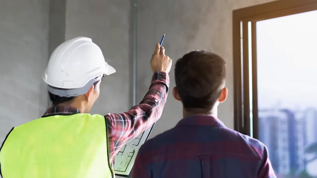 Inspector in a hard hat pointing at a window and interior wall during an inspection.