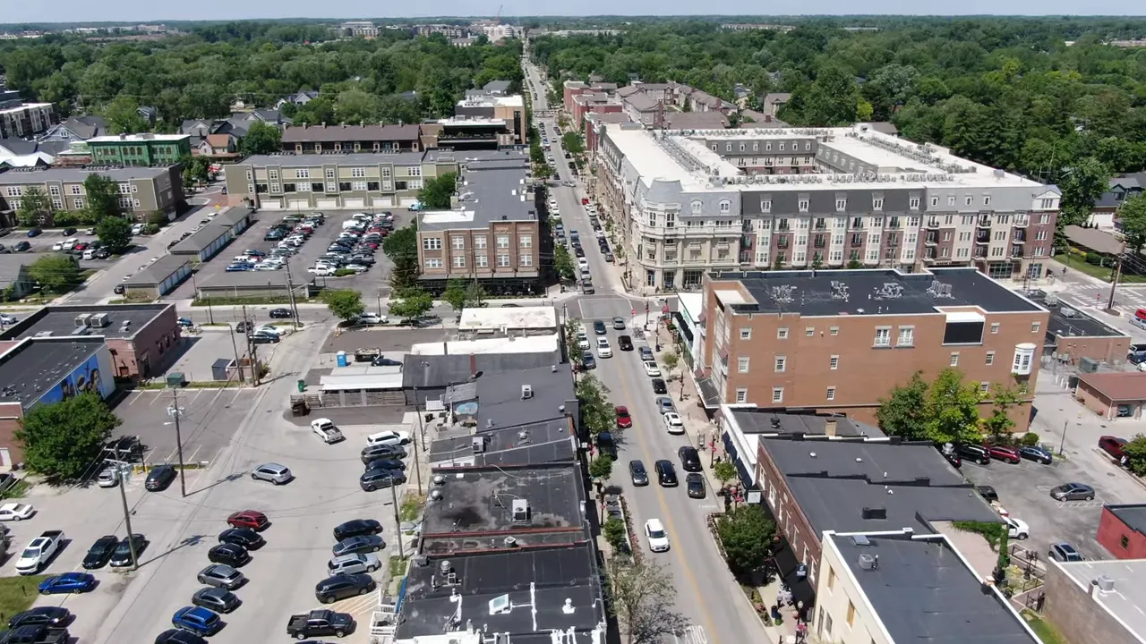 Aerial drone view of a downtown street with mixed-use buildings, parked cars, and tree-lined surroundings.
