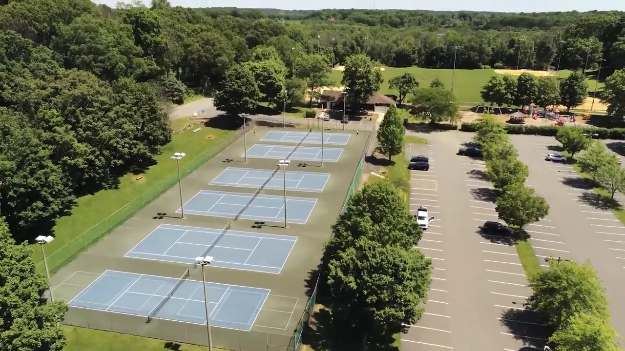 Aerial view of multiple outdoor tennis courts next to a large parking lot and trees
