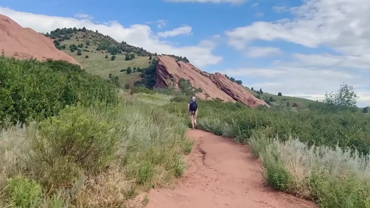 Person hiking along a dirt trail through green hills near red rock formations in Colorado