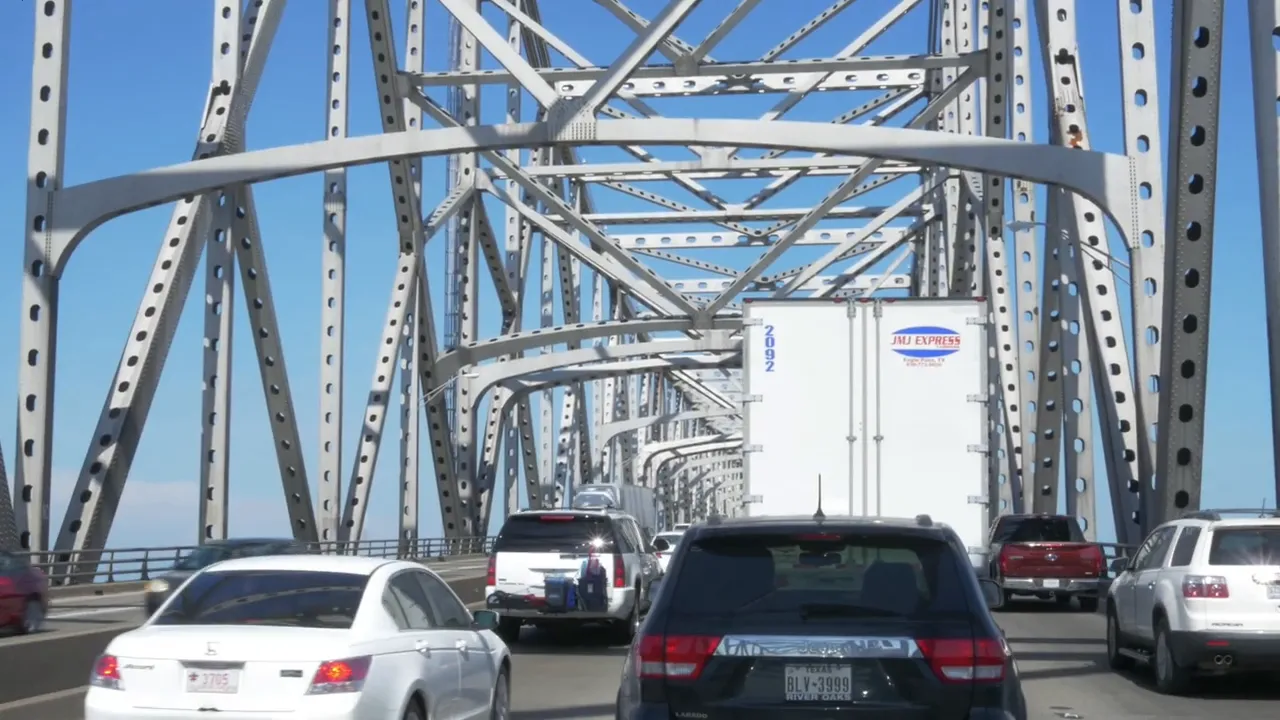 Vehicles and trucks in traffic crossing a steel truss bridge with clear daytime sky