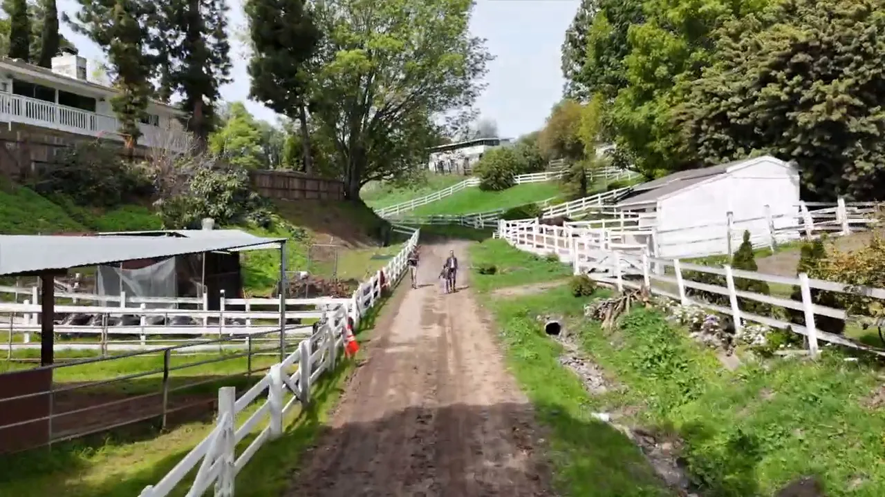 Dirt trail between white fences with grassy slopes and homes on both sides