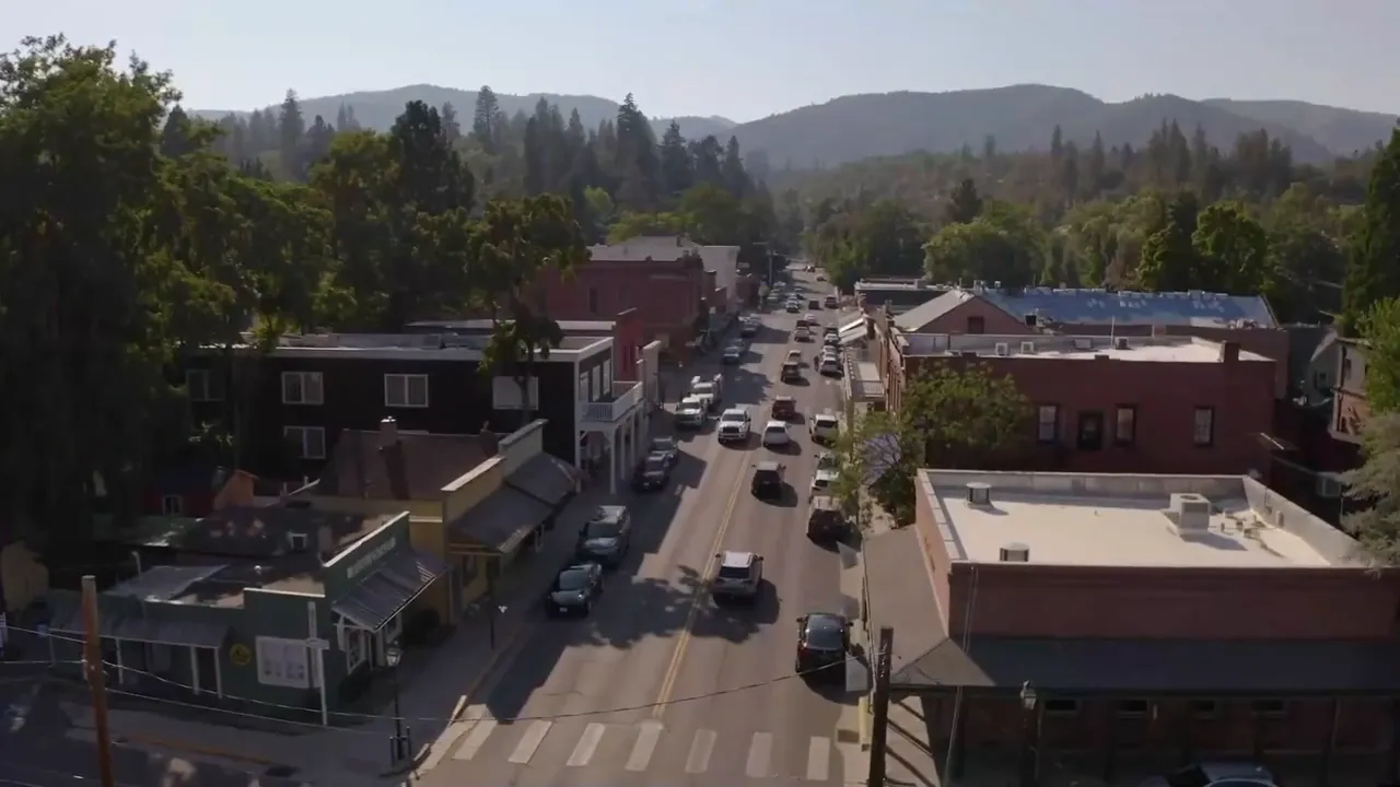 Aerial view down a downtown street in Jacksonville, Oregon with parked cars, brick buildings, and mountains in the background
