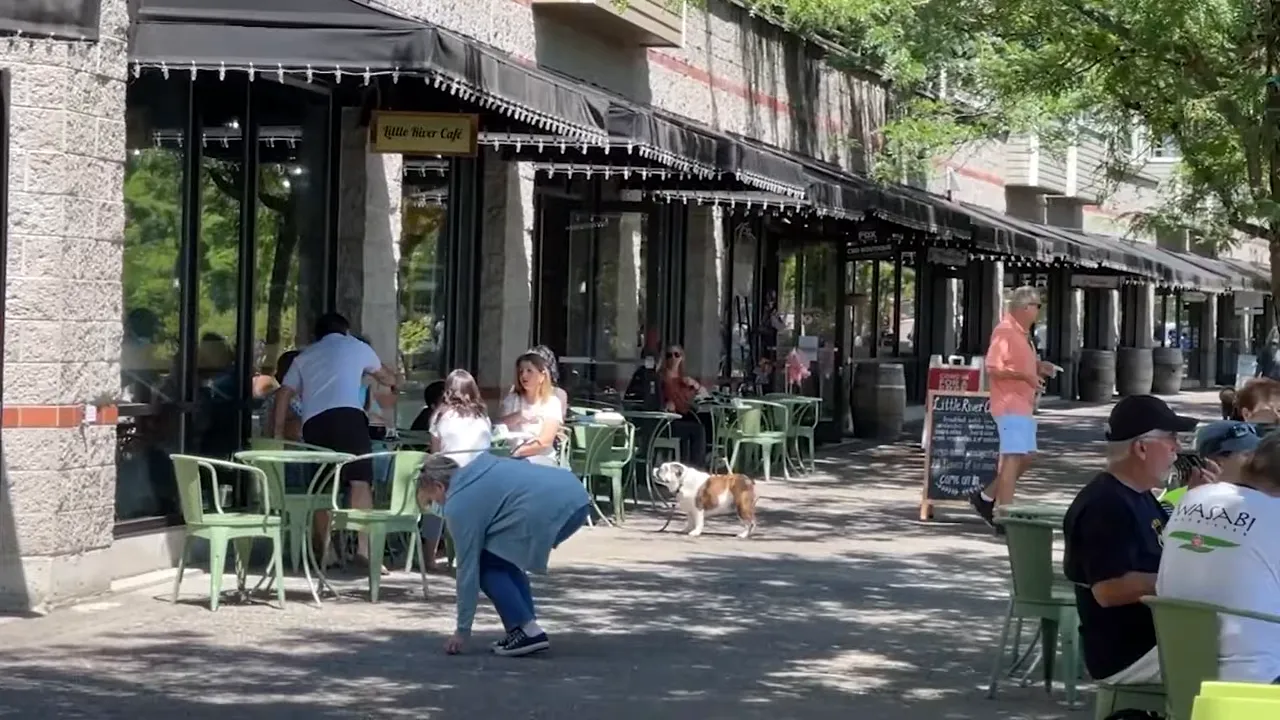 Outdoor cafe seating along a shaded sidewalk with people dining and walking past storefronts