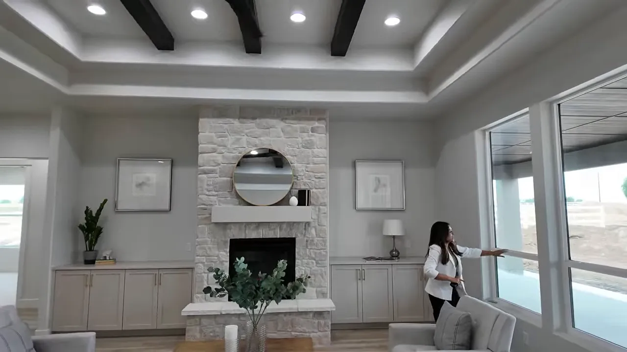 Open living room with tray ceiling beams ceiling fan stone fireplace and large windows