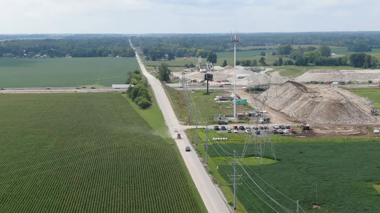 Aerial view of a rural road flanked by cornfields with a construction site and parked cars near Greenfield