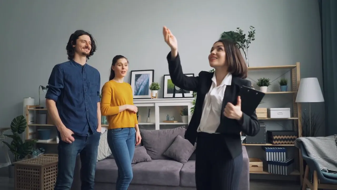 Realtor holding a clipboard showing a young couple around a home's living room during a walkthrough