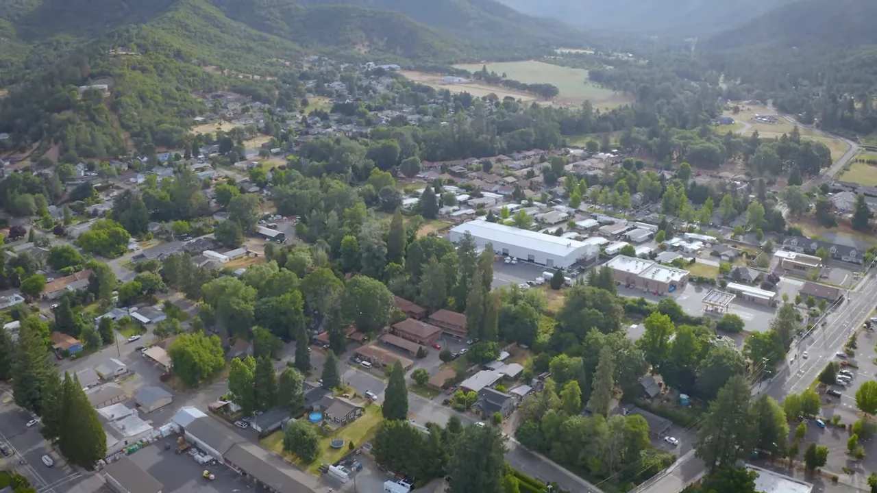 Wide aerial view of a small town with homes, commercial buildings, roads and surrounding forested hills