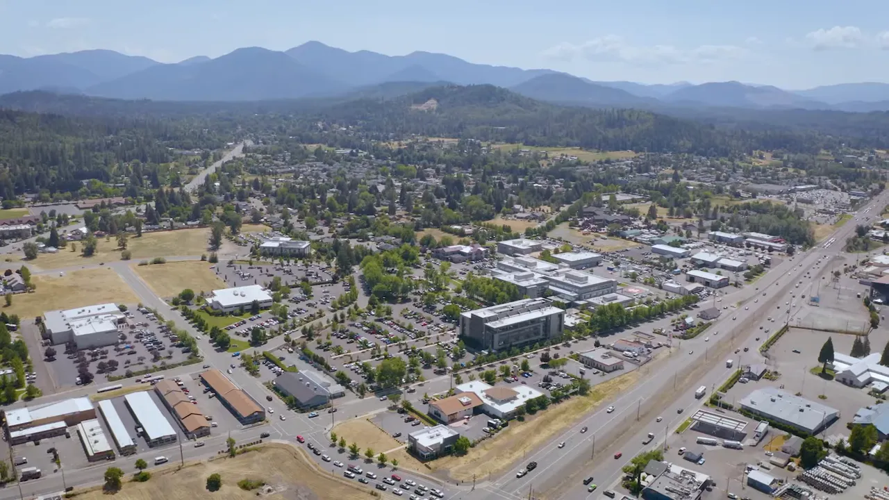 Wide aerial view of a small Southern Oregon town, parking lots and mountain backdrop.