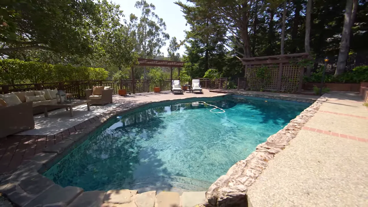 Bright, clear view of a rectangular backyard pool with deck seating, pergola and mature trees