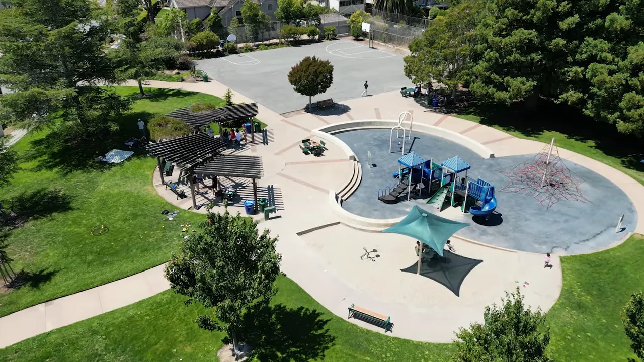 Aerial view of a playground with shade structures, picnic pergolas and surrounding grassy park area