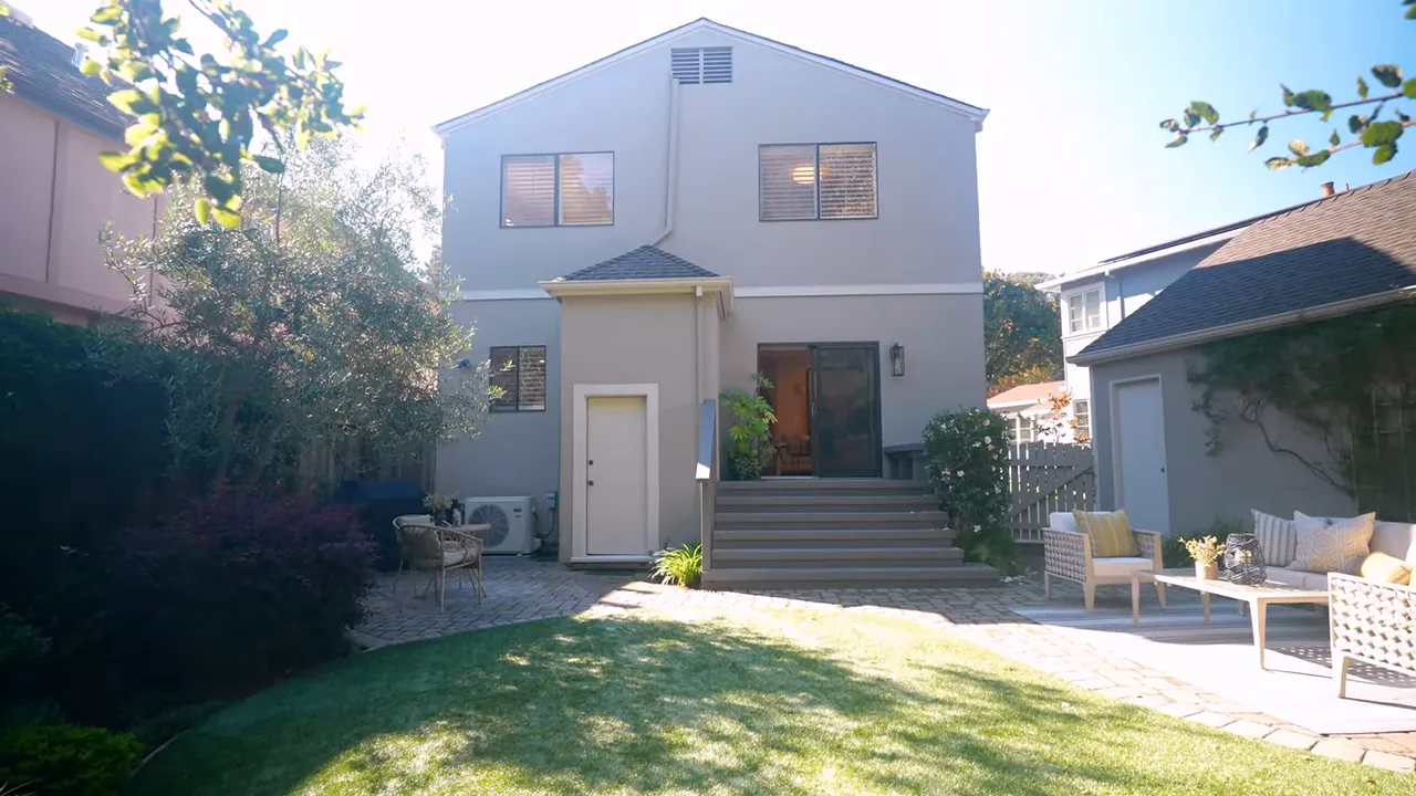 Backyard lawn, patio seating and two-story house exterior with trees