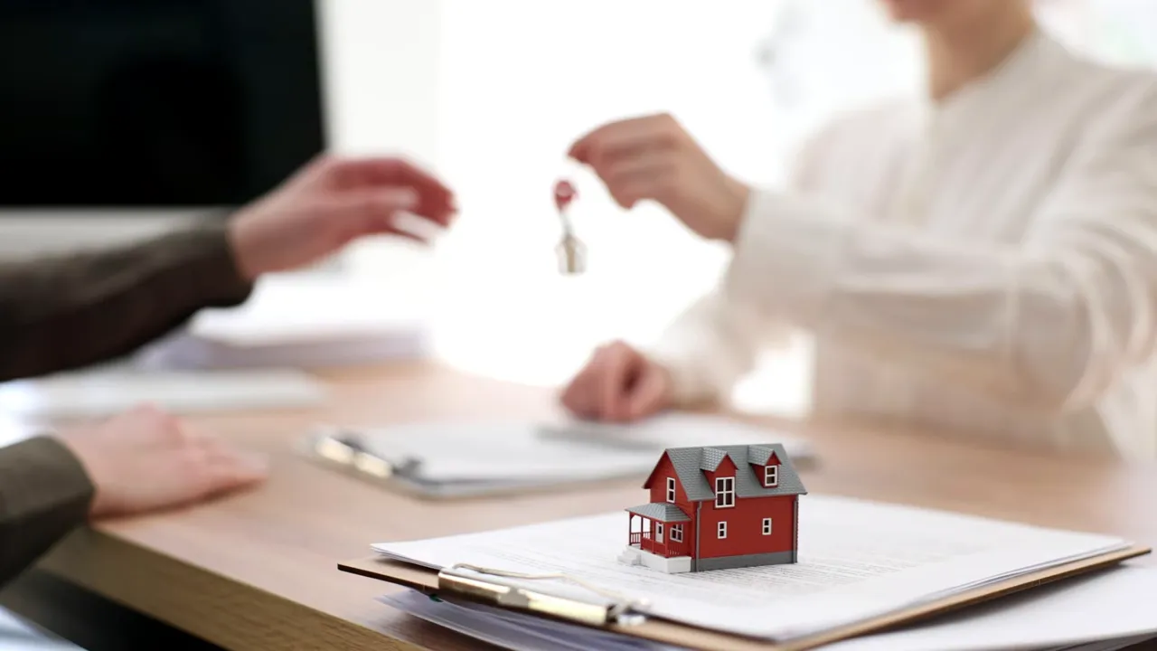 Person handing keys over above a small red model house sitting on contract paperwork