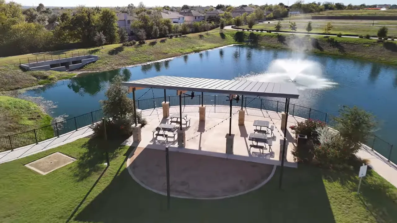 Aerial view of a covered pavilion with picnic tables beside a pond and fountain, with nearby walking paths and homes.