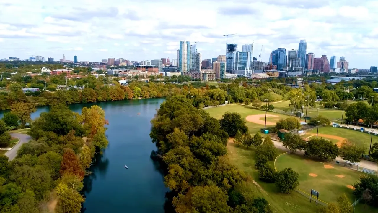 Aerial view of a tree-lined creek with adjacent ballfields and the Austin skyline — representing the Barton Creek Greenbelt.