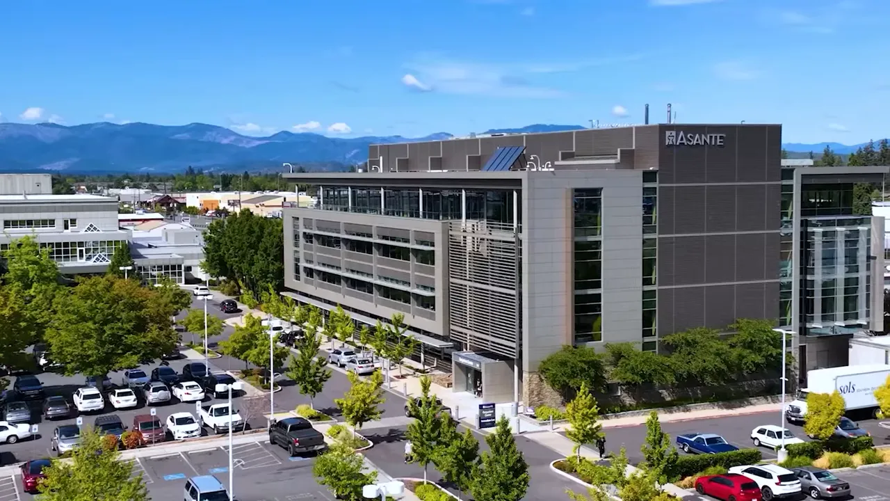 Aerial view of a regional hospital building (Asante) with parking, trees and mountains in the distance