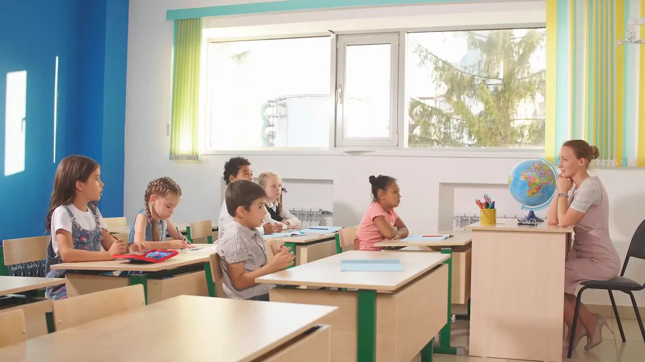Children seated in a classroom with a teacher at a desk