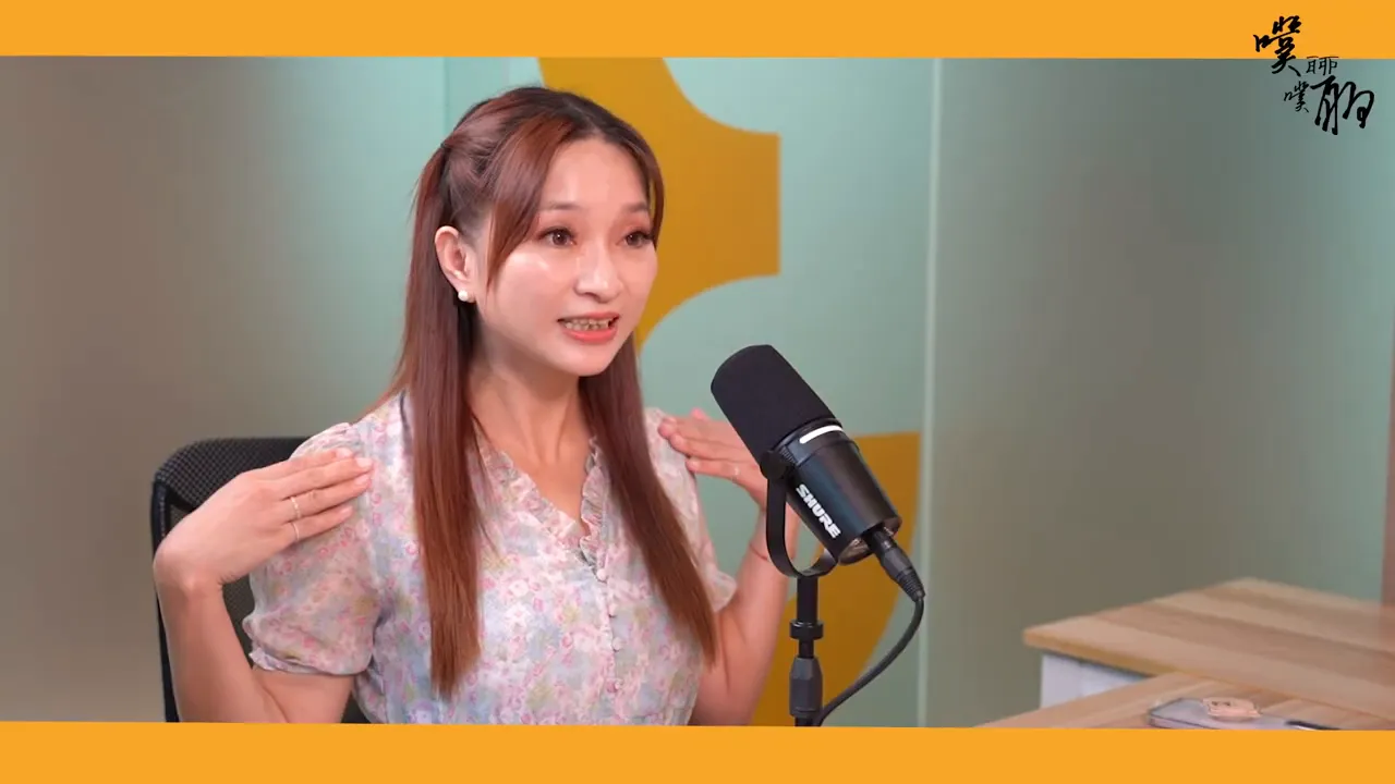 老師在麥克風前做出如同支援的手勢，雙手靠近肩膀，表情溫和且鼓勵。