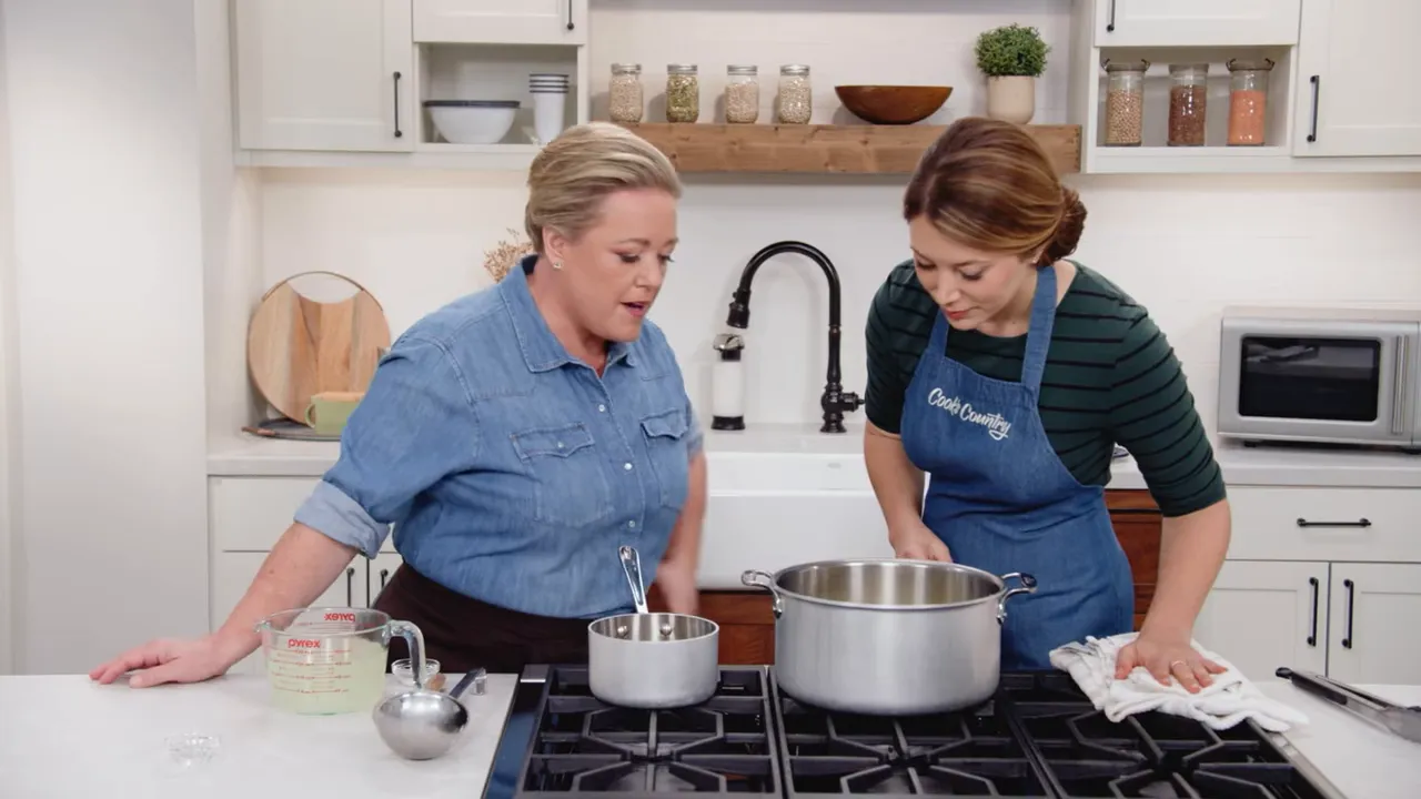 Two cooks leaning over a large pot on the stove, finishing spaghetti with reserved pasta water nearby