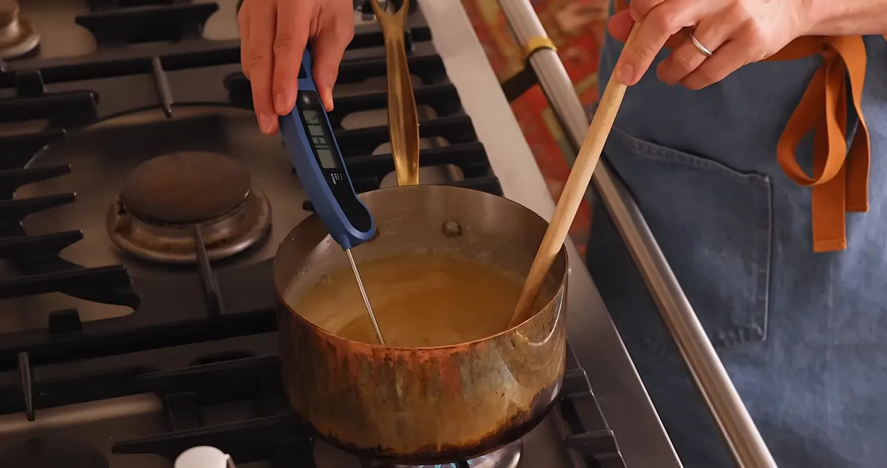 Hands holding a candy thermometer and wooden spoon while checking caramel temperature on the stove