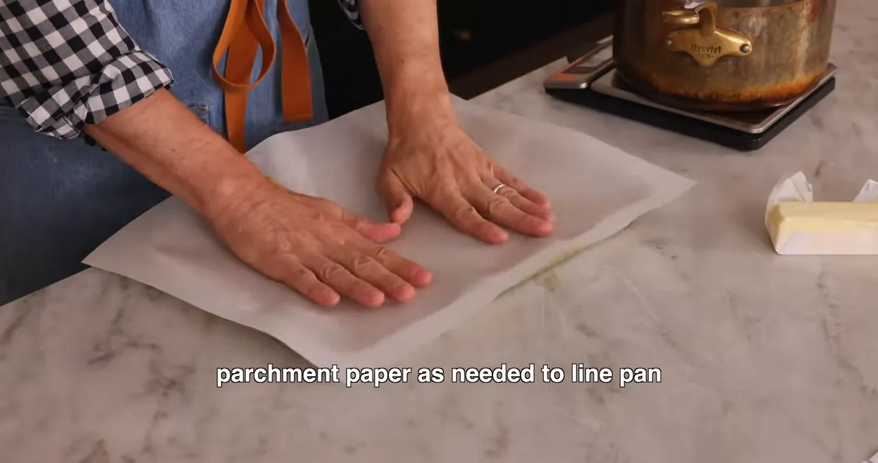Hands smoothing parchment paper into a baking pan with a pot and butter nearby