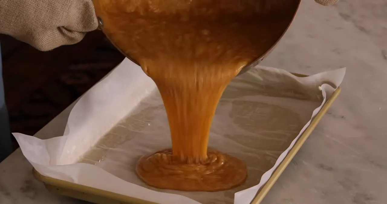 Golden caramel being poured from a saucepan into a parchment-lined baking pan, forming a thick stream and pooling in the center.