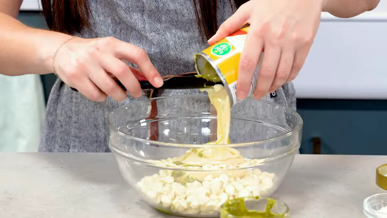 Pouring sweetened condensed milk into a glass bowl with white chocolate chips and pistachio cream, spatula ready.