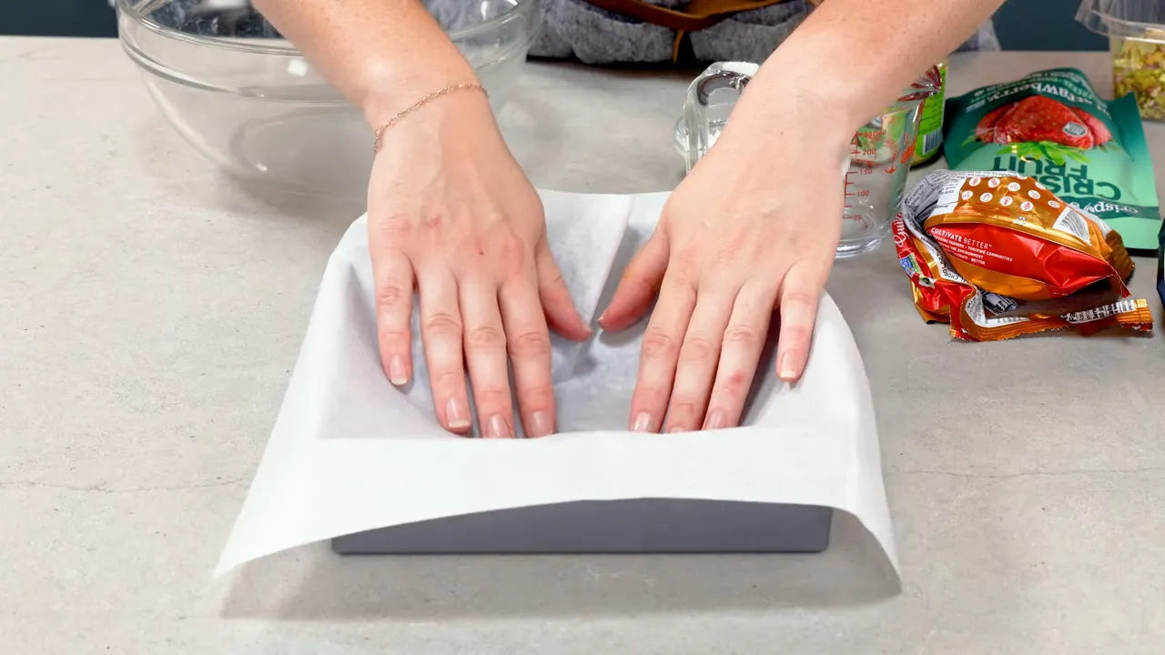 Hands pressing parchment paper into a lined 9x9-inch baking pan on a countertop.