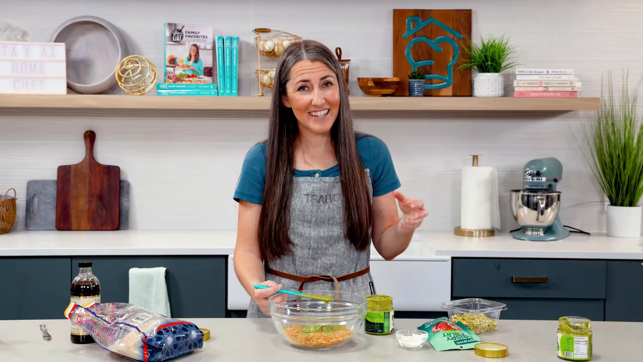 Clear bowl with toasted kataifi and a spatula adding green pistachio cream beside other ingredients on the counter