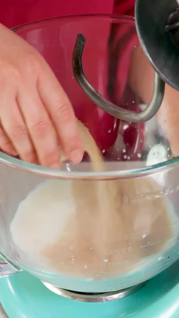 Hand sprinkling yeast and sugar into a glass mixing bowl with a dough hook visible