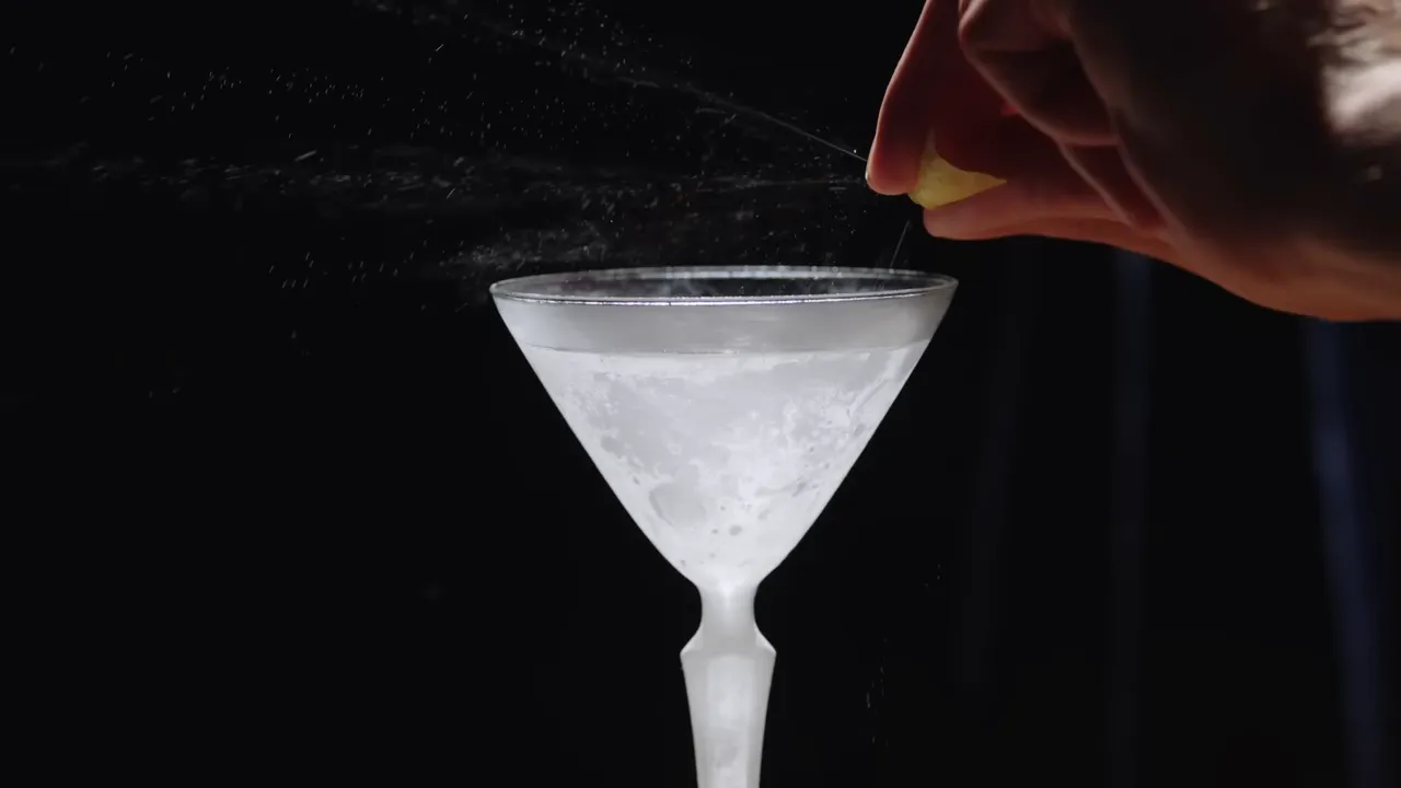 Hand squeezing a lemon peel over a martini glass with a visible spray of citrus oil
