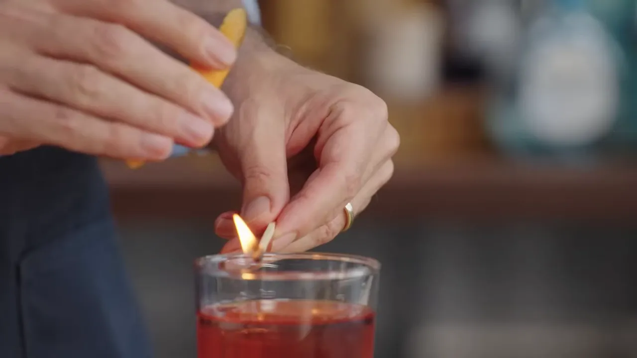 Hand holding an orange peel over a match flame above a cocktail to express citrus oils