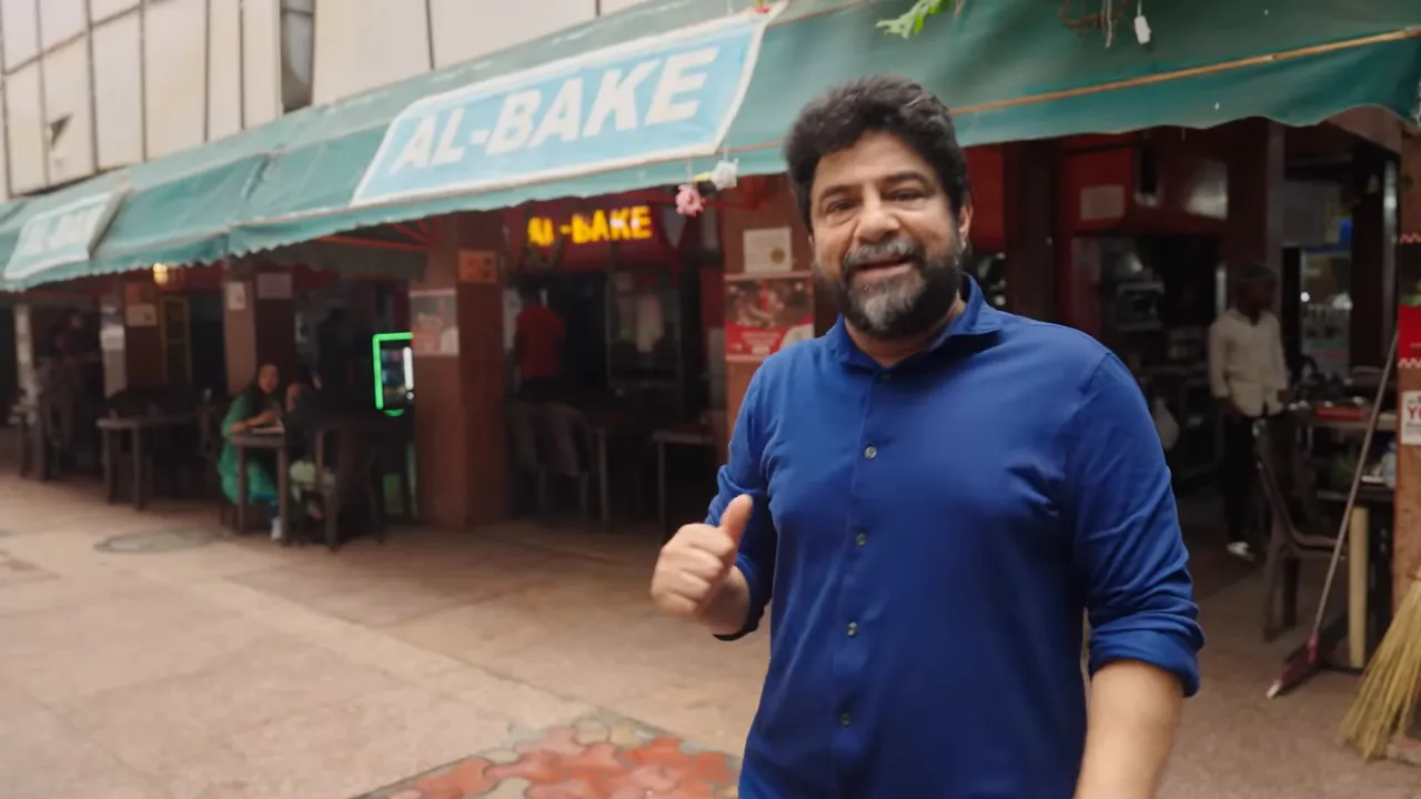 Presenter smiling in front of the Al‑Bake storefront sign and entrance.