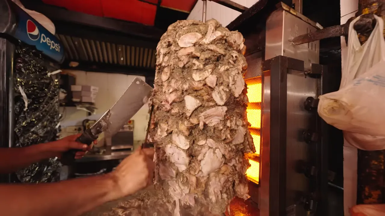 Large marinated shawarma stack being sliced beside glowing burners