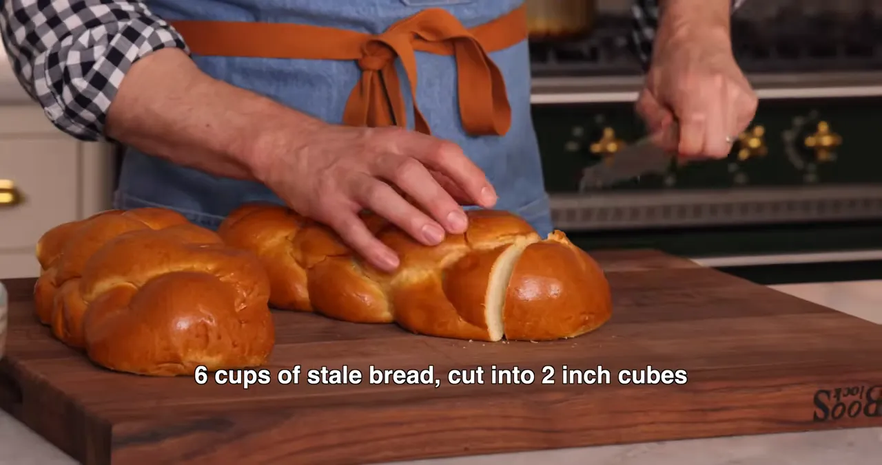 Close-up of hands slicing a brioche loaf on a wooden cutting board with on-screen text: '6 cups of stale bread, cut into 2 inch cubes'.