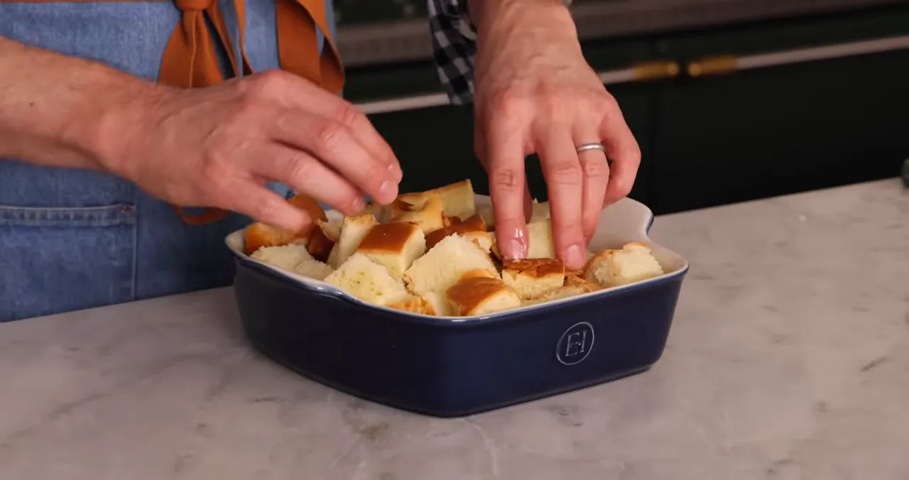 hands arranging cubed brioche in a blue baking dish, tucking crust pieces up