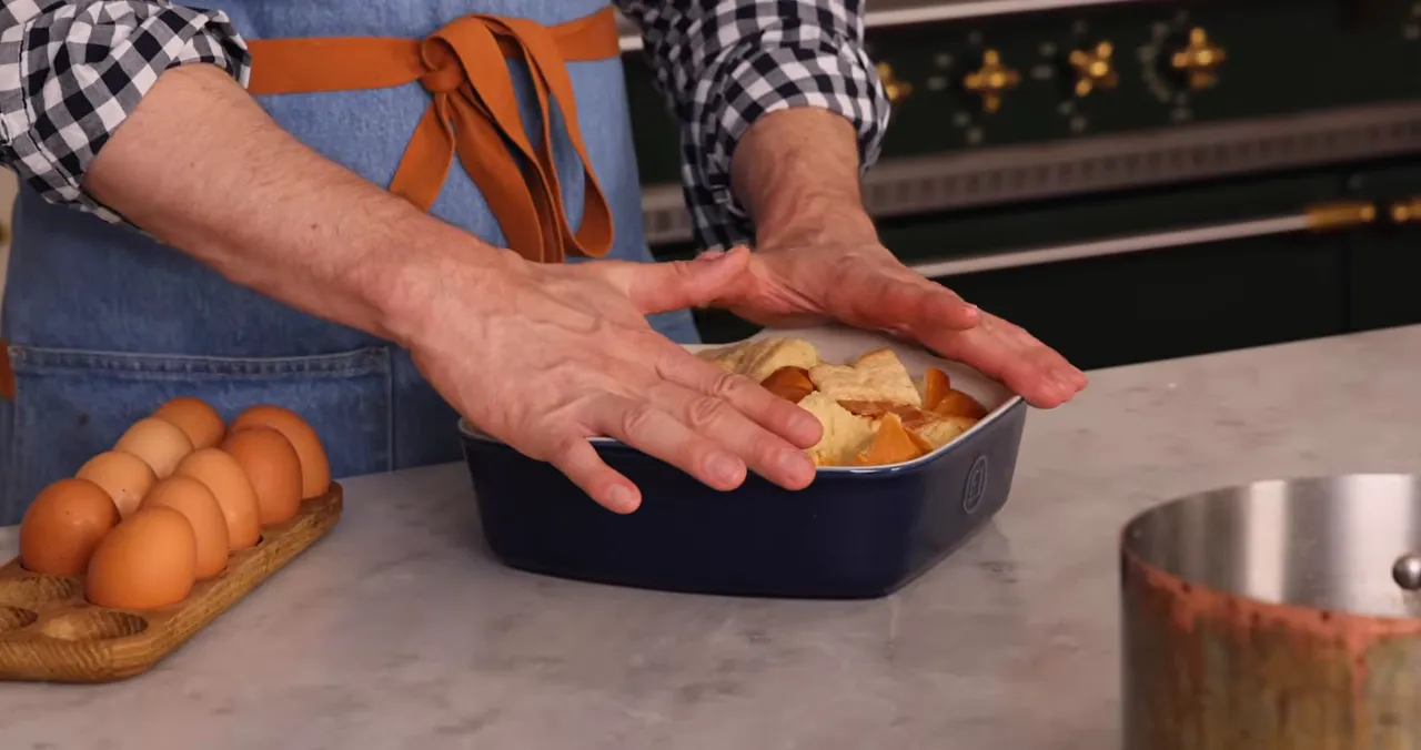 Close-up of hands pressing cubed bread in a baking dish next to a row of eggs and a saucepan