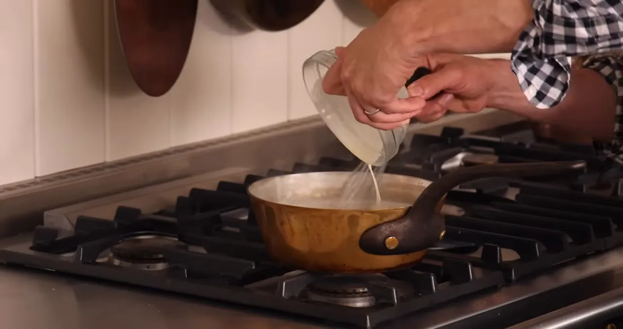 Hands pouring a cornstarch slurry from a small bowl into a copper saucepan of milk on the stove.